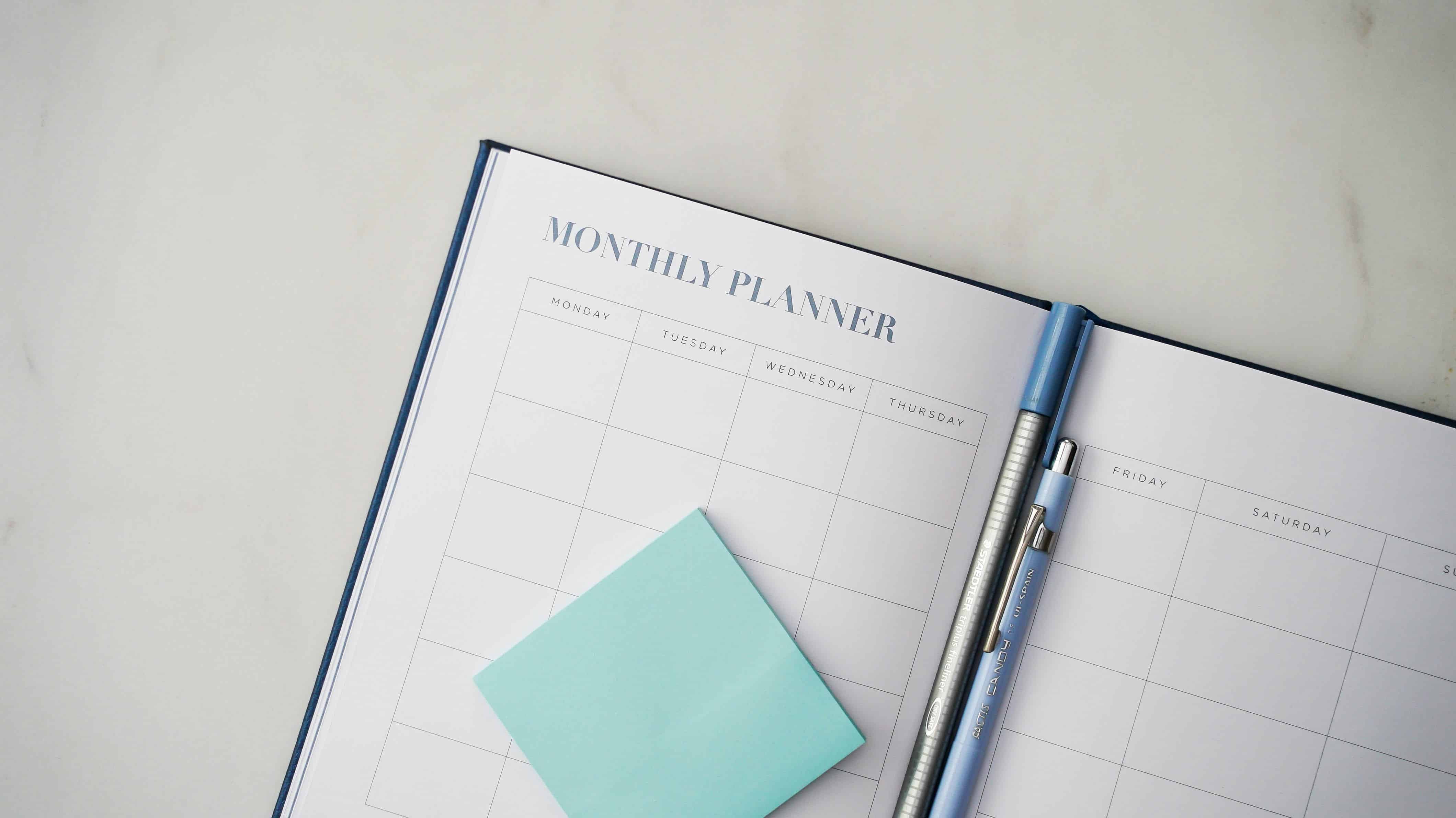 Empty planner, open at a month with pens and a post it note pad showing that something needs to be planned